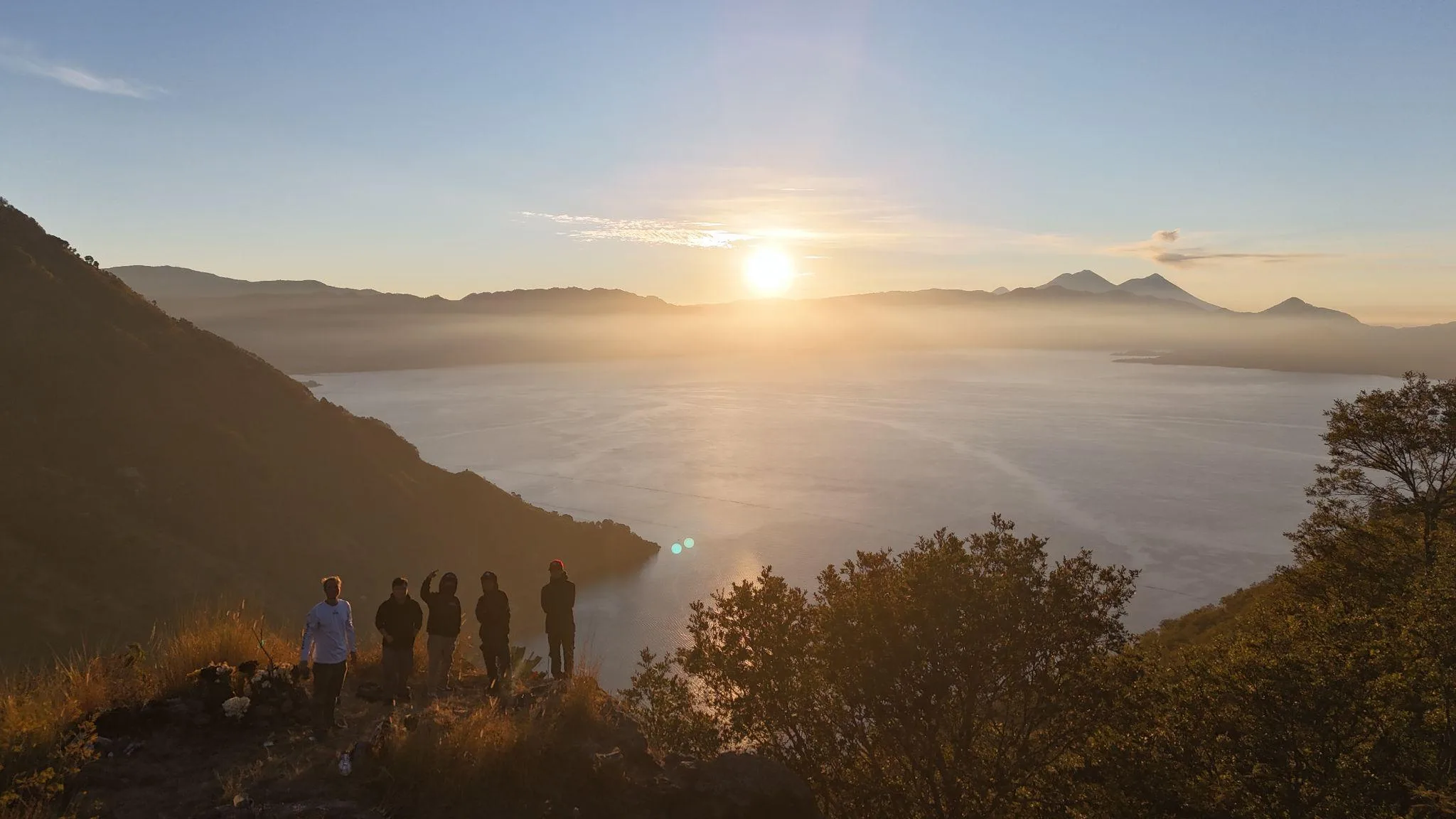Sunrise over Lake Atitlán from a ridge above Tzununá, hikers silhouetted against the water with the volcanoes rising on the far shore